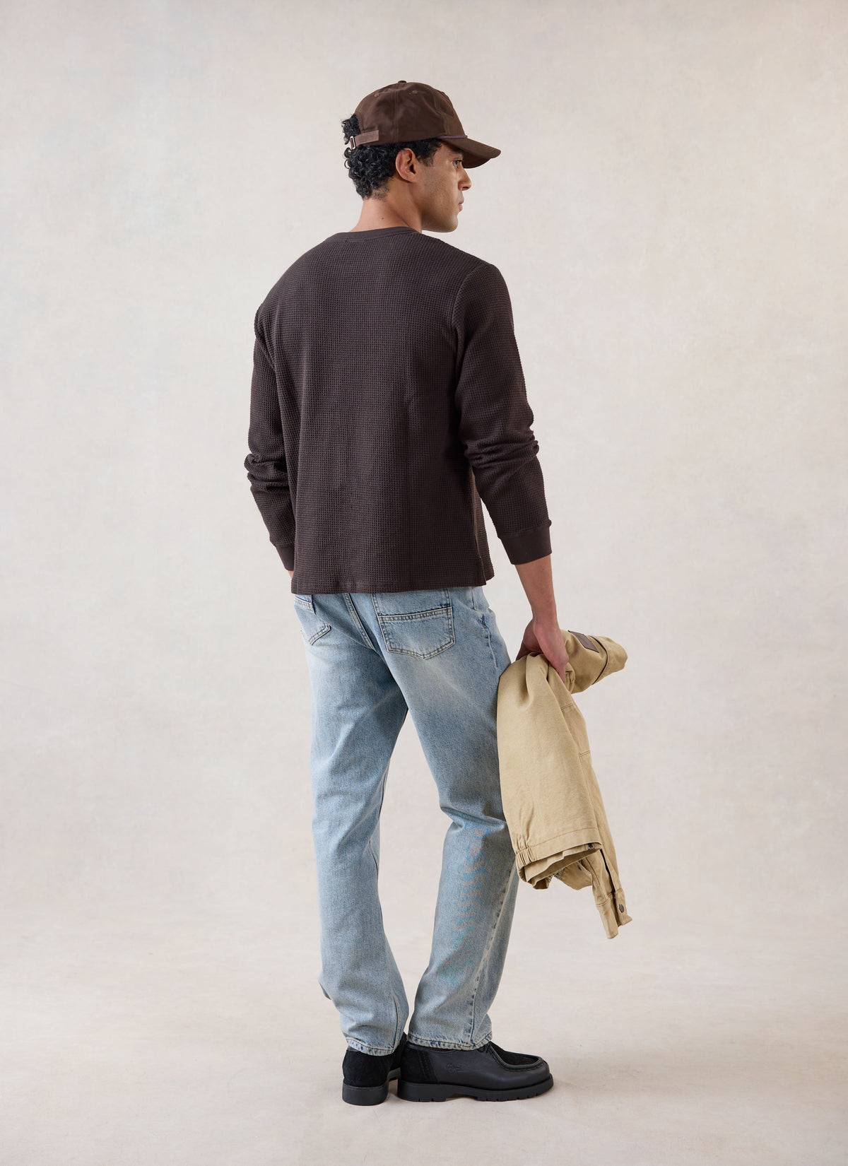 A model from the back wearing the brown Percival waffle long sleeve T-shirt, light blue jeans, cap, and holding a jacket.