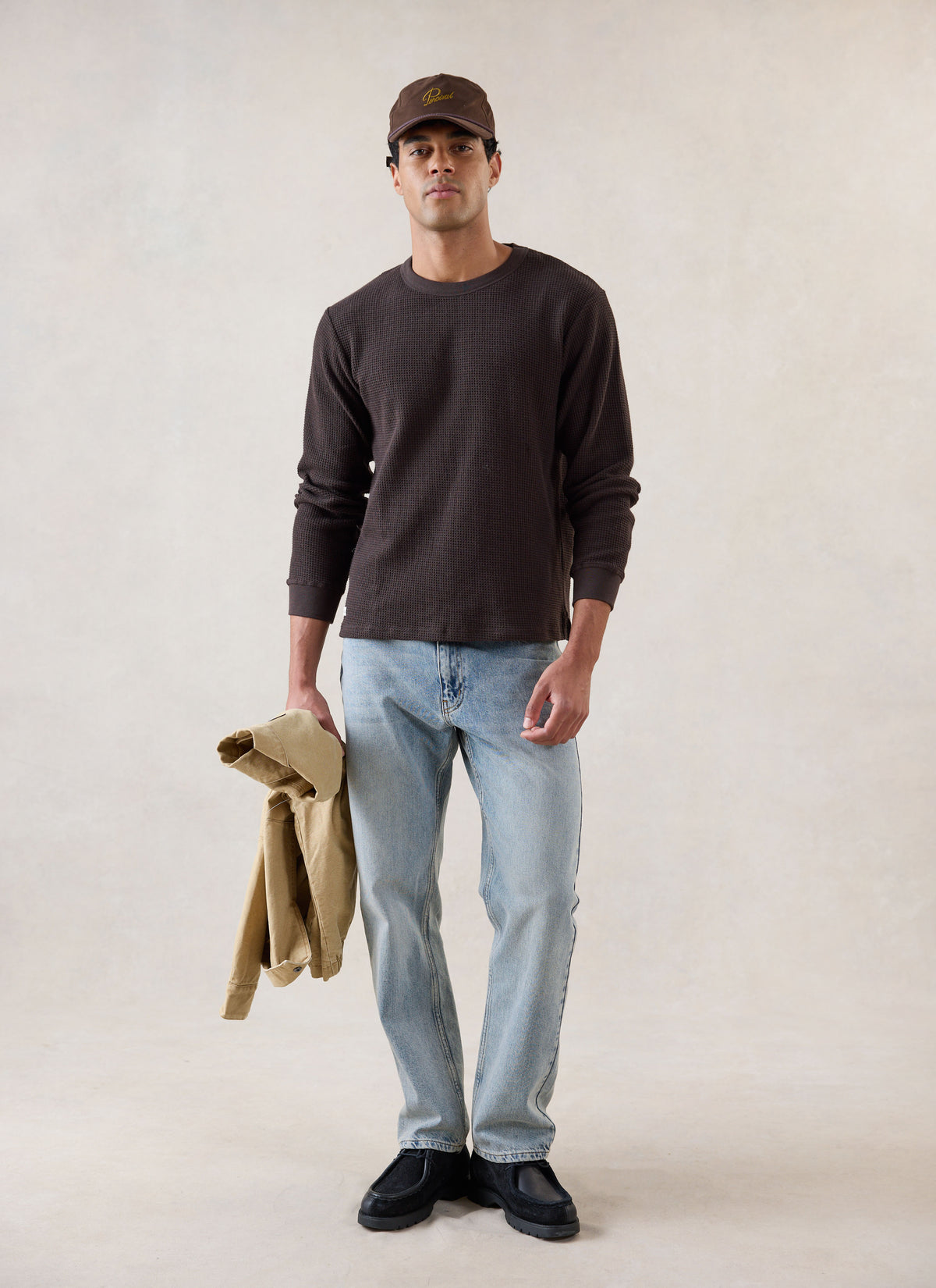 A model wearing the Percival brown waffle long sleeve T-shirt with blue jeans, a cap, and holding a jacket.