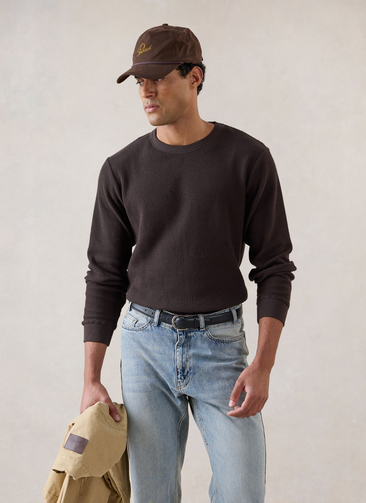 A model wearing the Percival brown waffle long sleeve T-shirt in 100% cotton with light blue jeans and a brown cap.