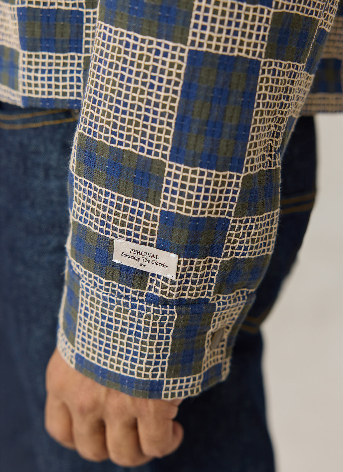 A close-up of the Percival Wallis grid check overshirt in navy blue and cream cotton, showing the branded cuff label.