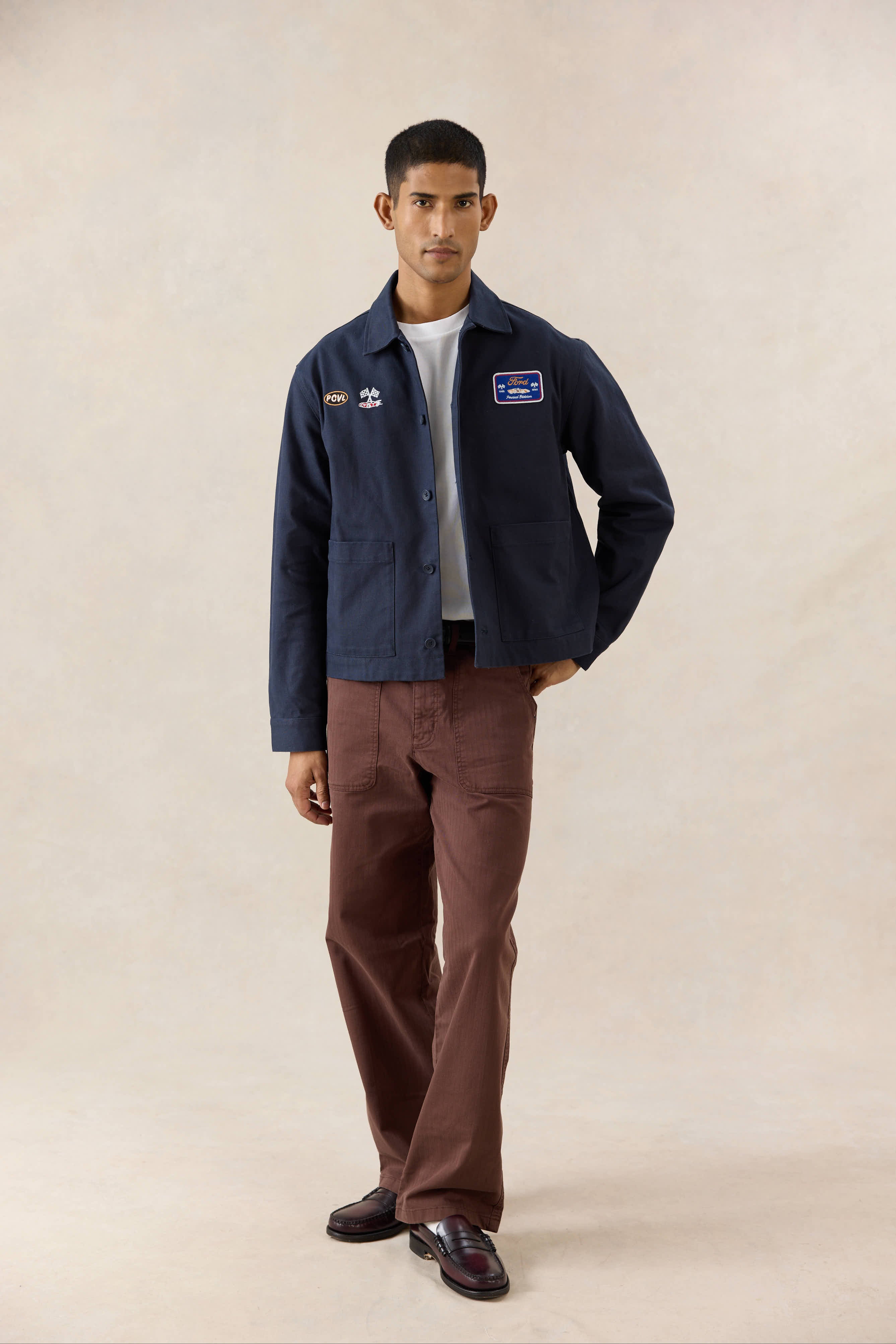 Navy Percival x Ford GT40 canvas jacket, paired with a white t-shirt and straight-leg brown trousers in a studio shot.