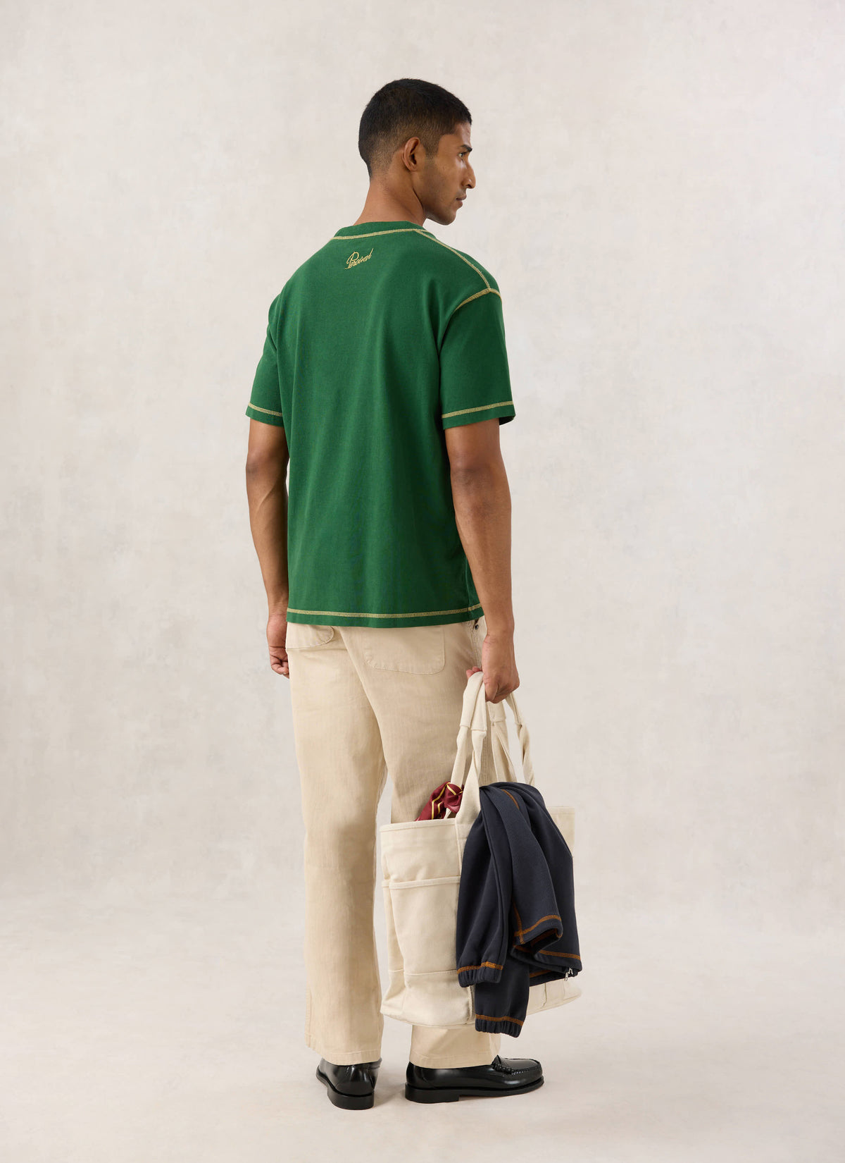Rear view of a model wearing the forest green Vintage Wash Oversized T Shirt by Percival x Guinness, holding a tote bag.