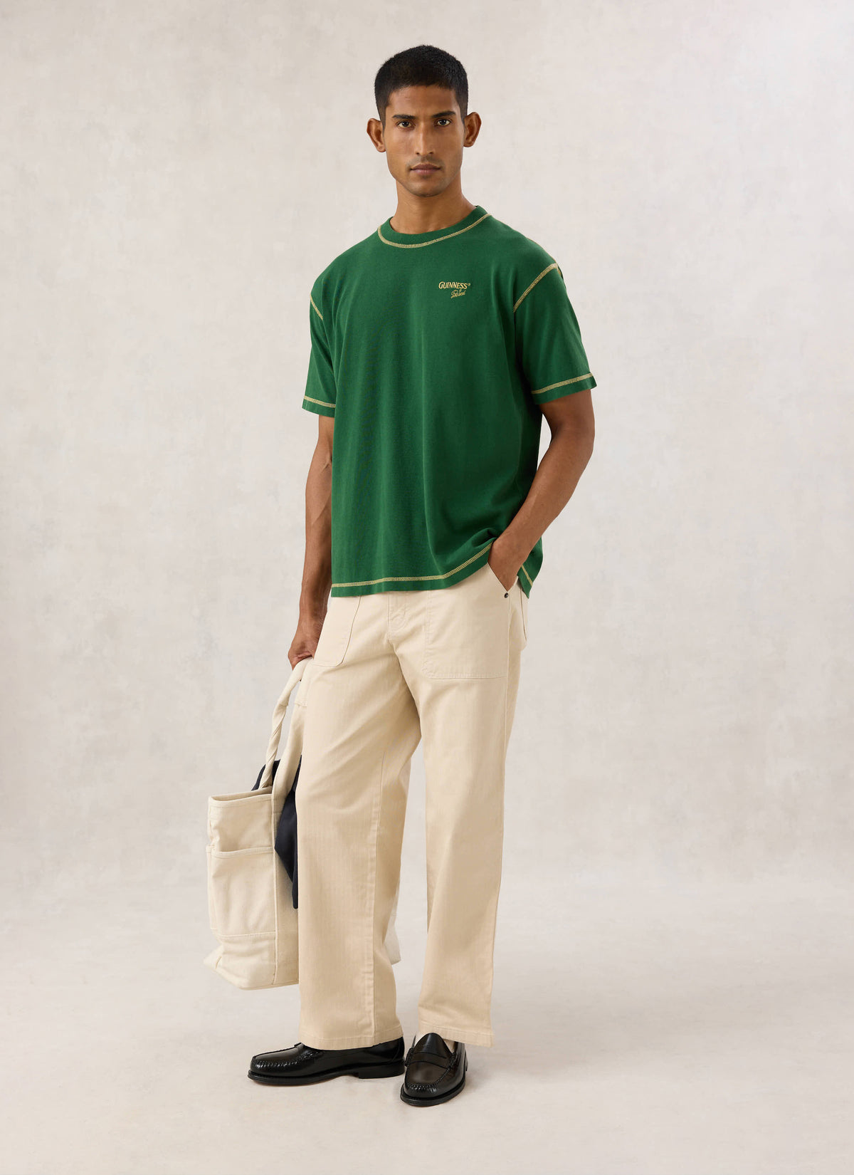 Model wearing the forest green Guinness x Percival T-shirt, paired with light trousers and holding a tote bag.