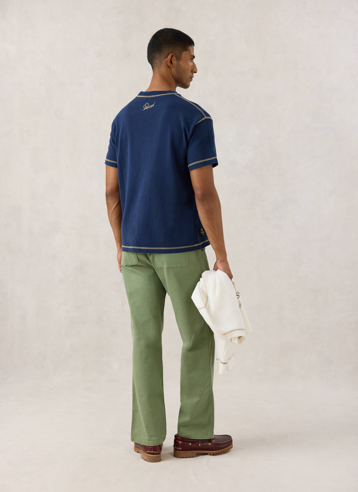 Rear view of a model wearing the navy Guinness x Percival T-shirt, showing the back neck embroidery and green trousers.