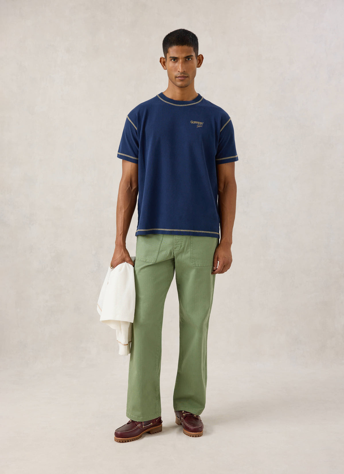 Model wearing the navy Guinness x Percival organic cotton oversized T-shirt with green trousers and holding a white layer.