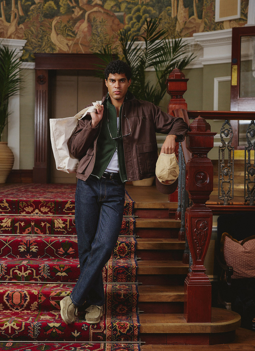 A model wearing a Percival espresso wax zip jacket over a forest green zip cardigan, raw jeans and wallabees on stairs.