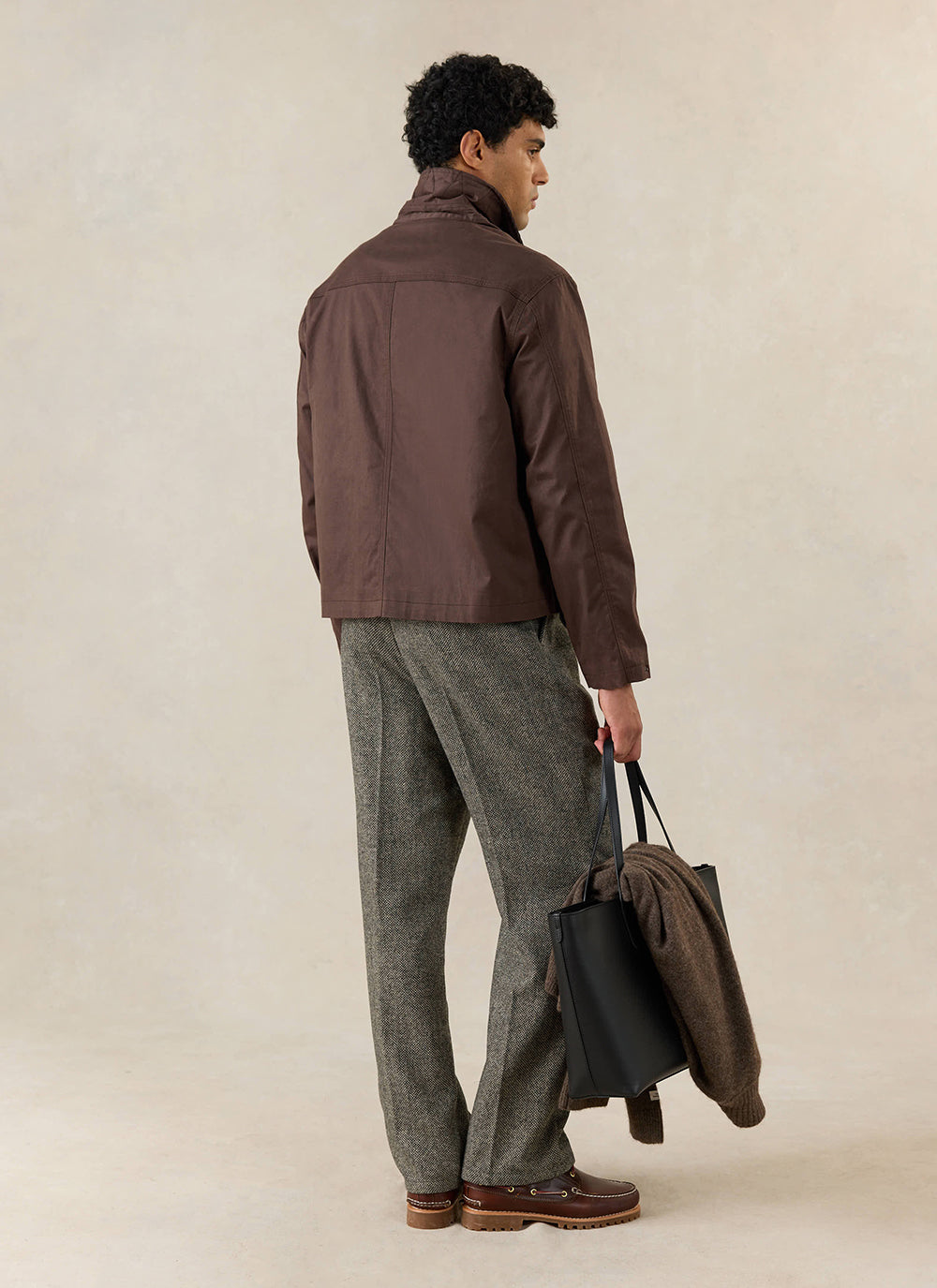 A model from the back wearing the Percival wax zip jacket in espresso, with grey trousers and a black tote bag.