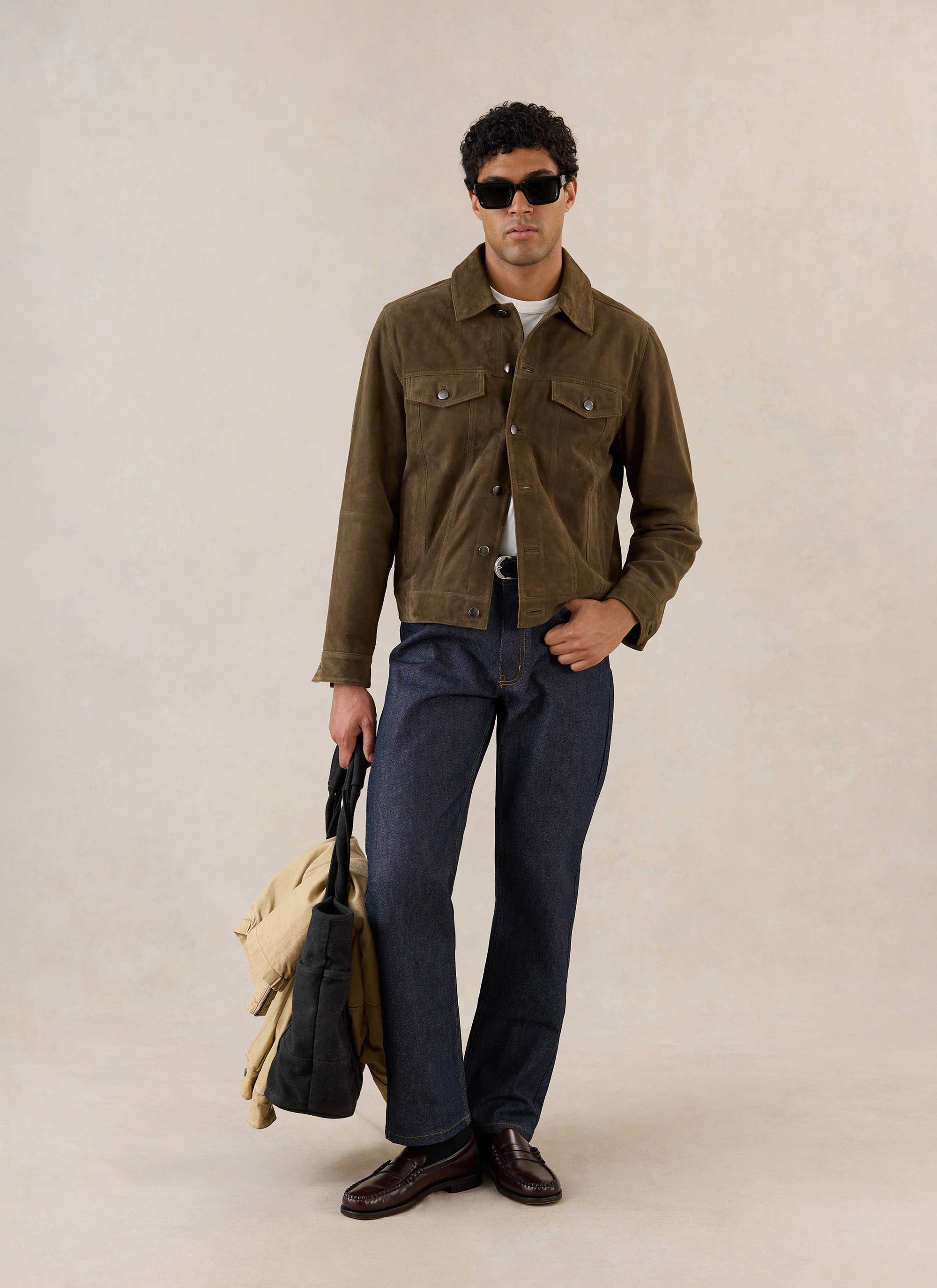 A model wears the olive Percival suede western jacket, styled with dark jeans and holding a tote bag in a studio.