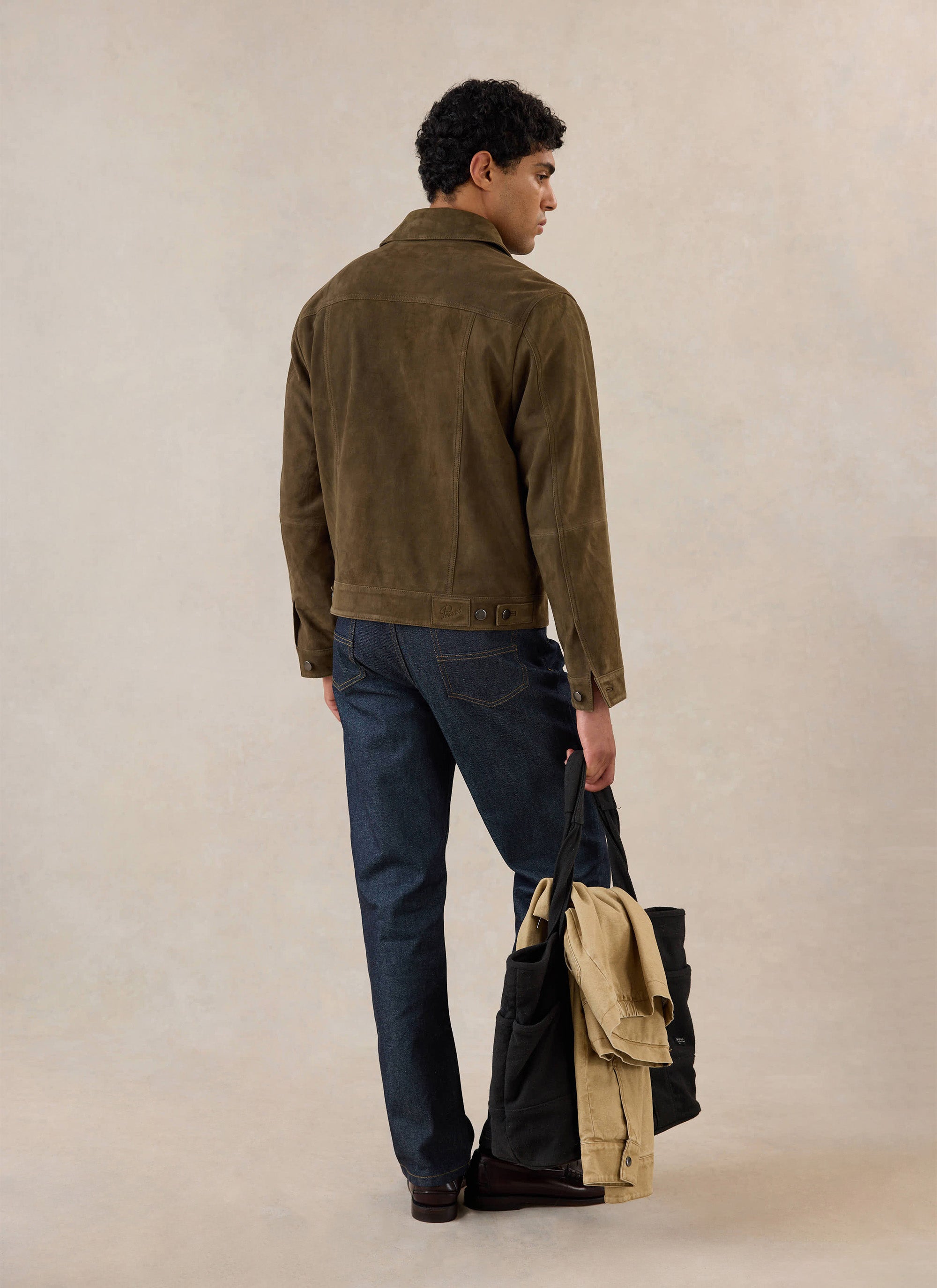 Rear view of a model wearing the olive suede Western Jacket from Percival, styled with dark jeans and holding a tote bag.