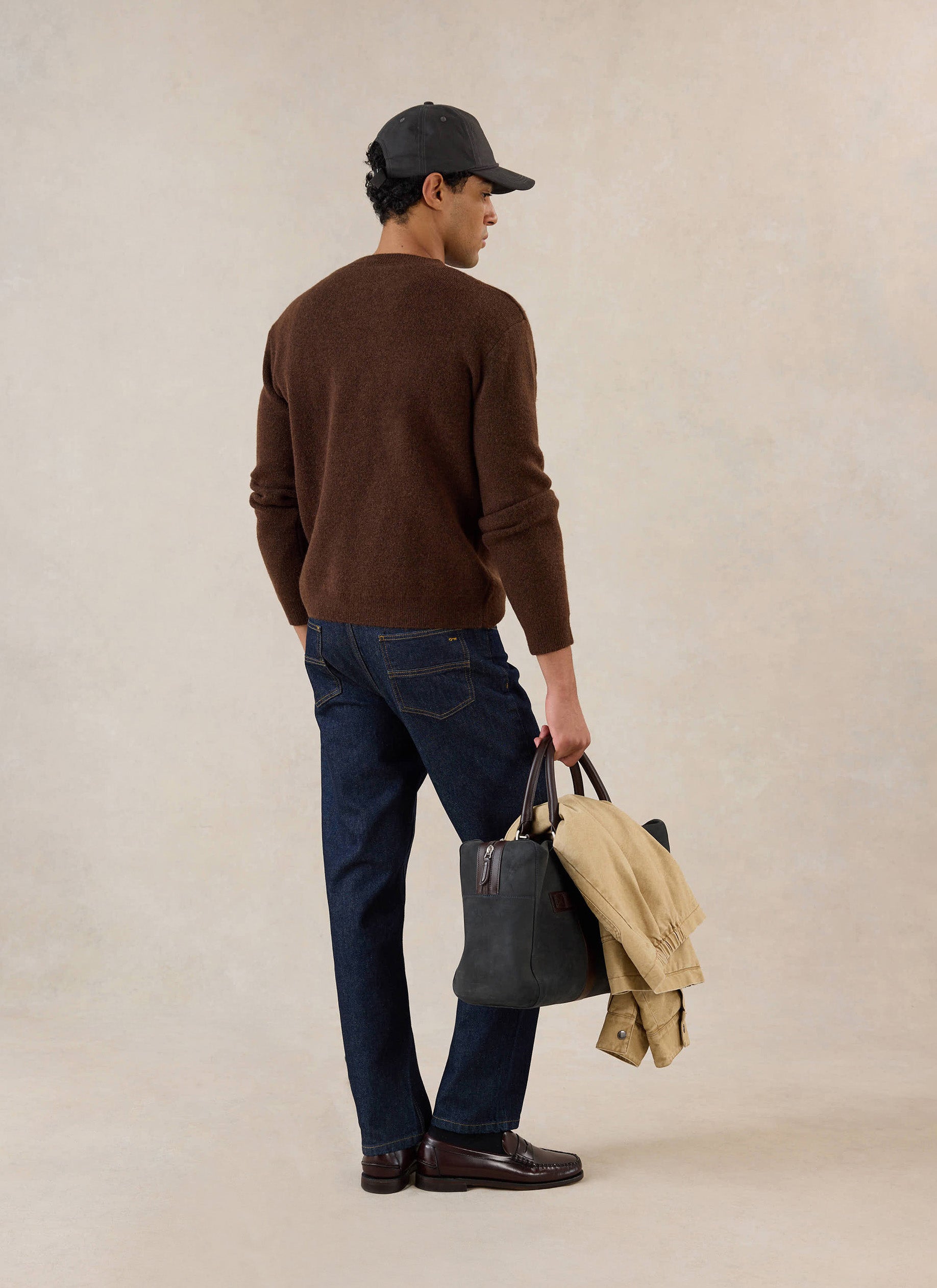 A model from the back wearing the espresso Percival mohair jumper, dark jeans, cap, and holding a bag and jacket.