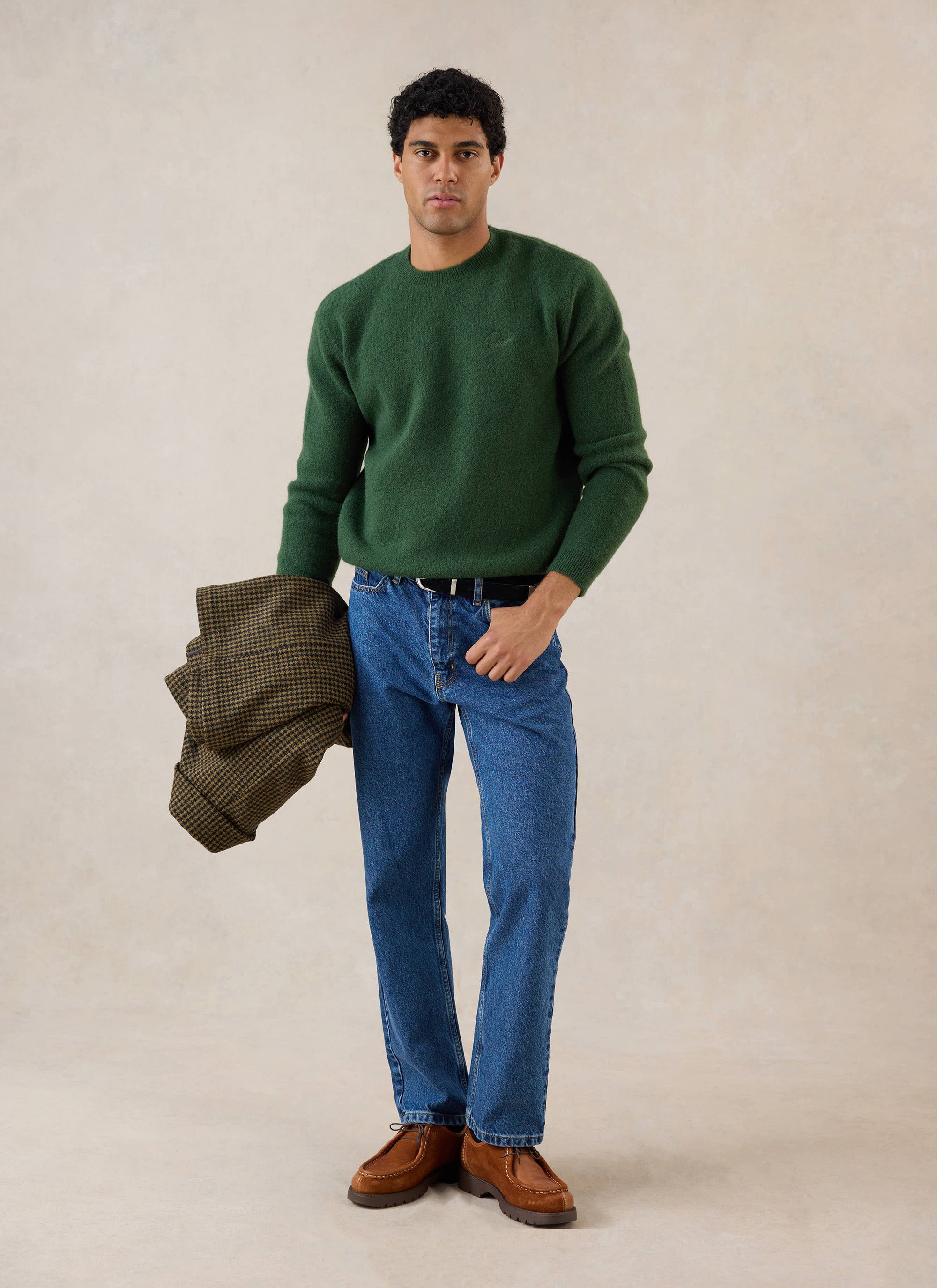 A model wears the forest Percival White Glove Boxy Crew Neck Jumper, styled with blue jeans and holding a patterned jacket.