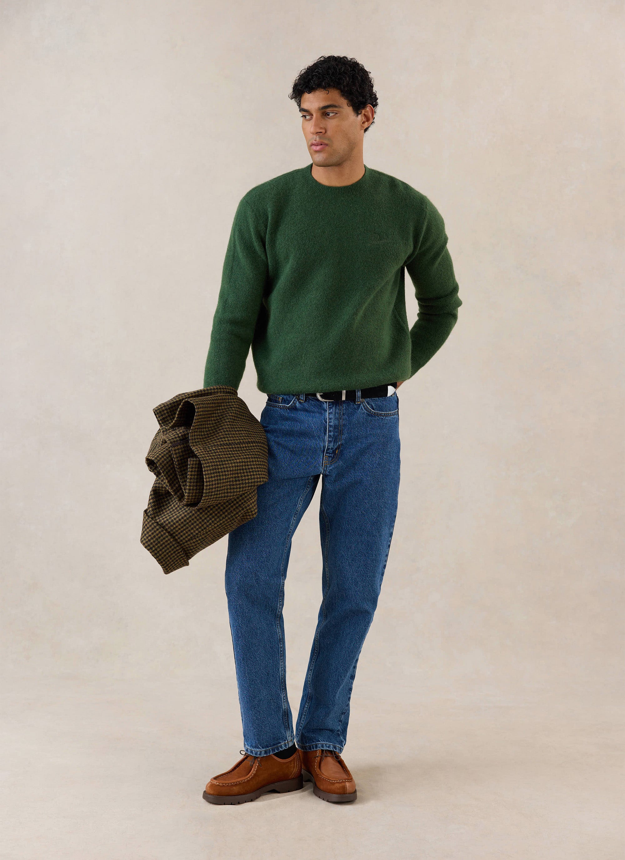 A model wears the forest green Percival mohair crew neck jumper with blue jeans and brown shoes, holding a patterned jacket.