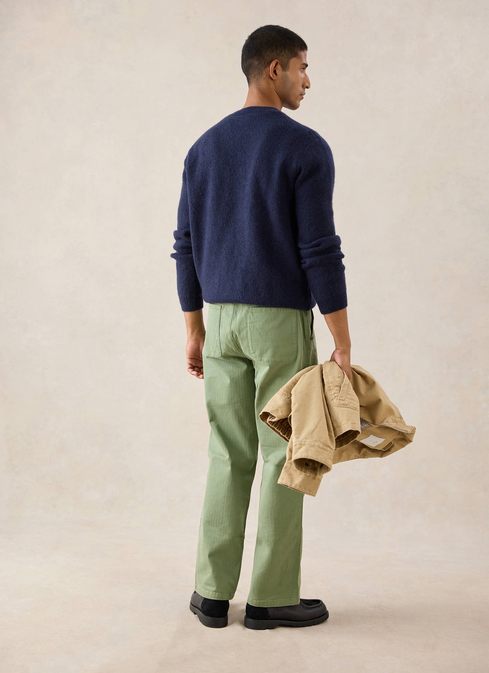A model wears the navy Percival jumper from behind, holding a tan jacket and wearing green trousers.