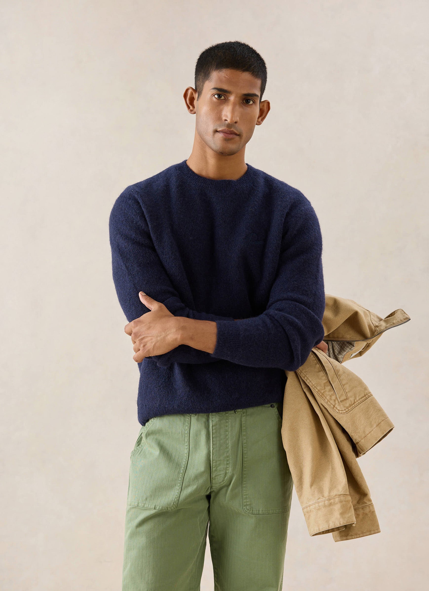 A model wears the navy Percival mohair crew neck jumper with green trousers, holding a beige jacket over his arm in a studio.