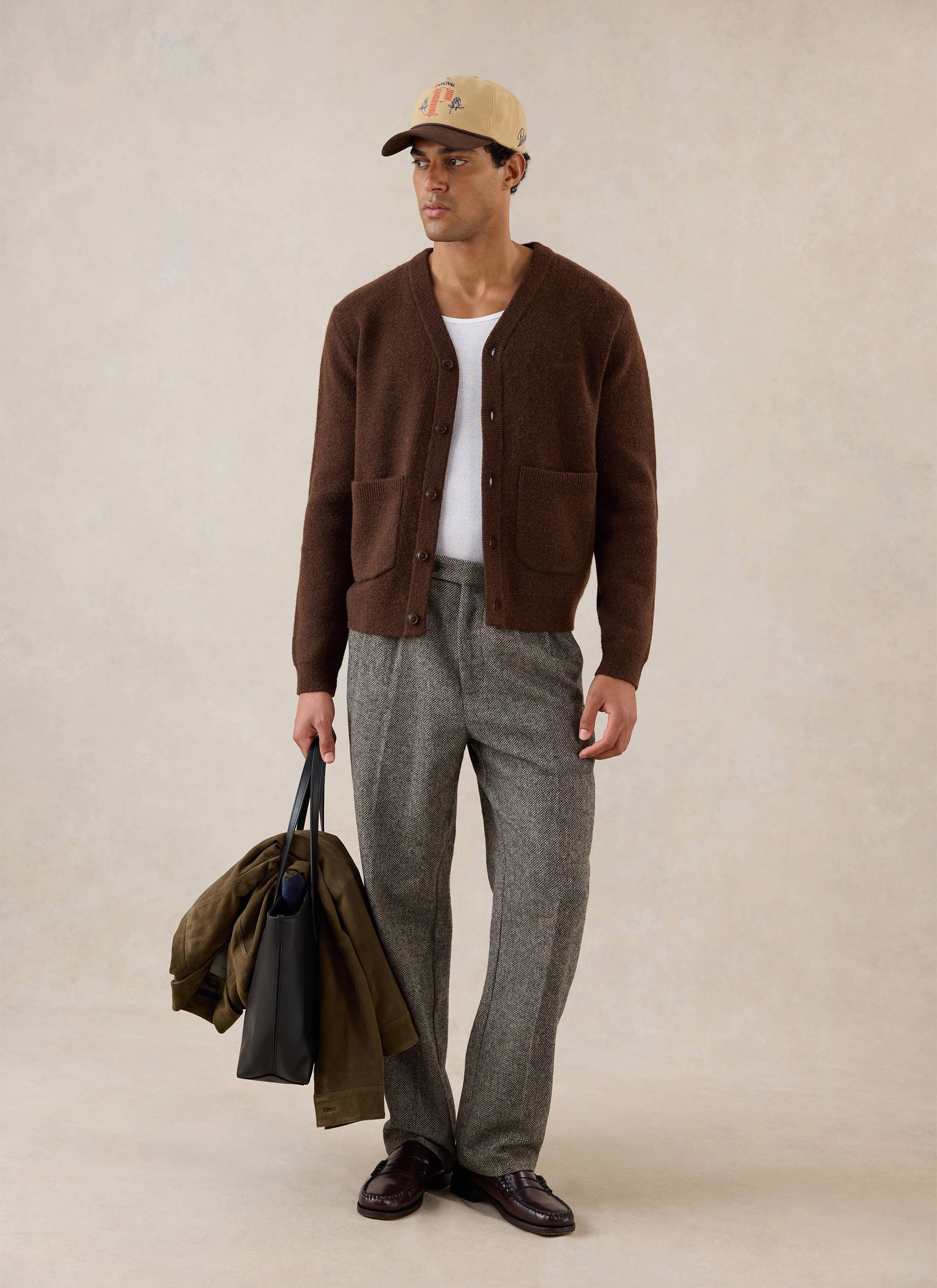 A model wearing the Percival espresso brown White Glove cardigan, styled with trousers, a cap, and a tote bag.