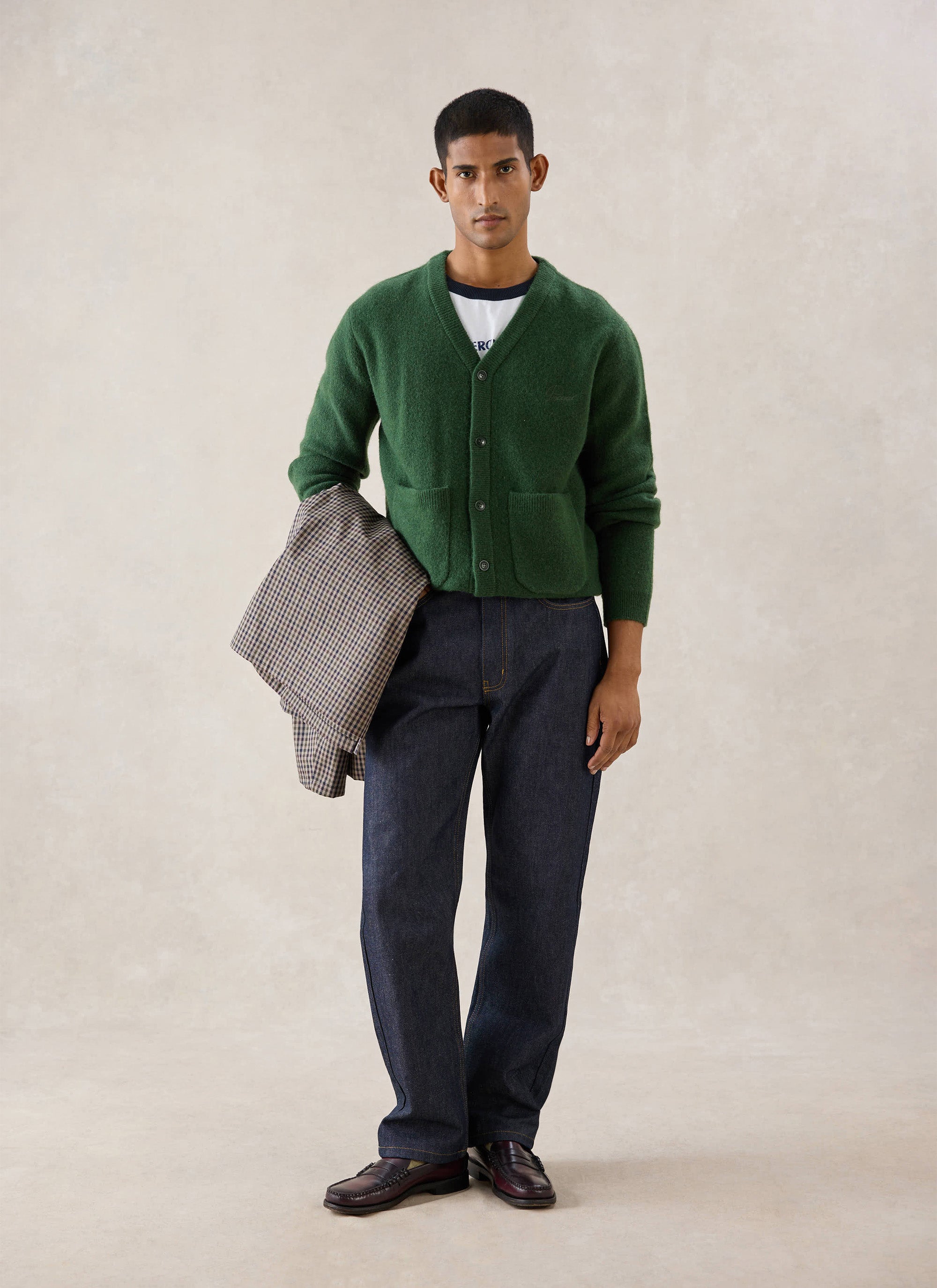 A model wearing the Percival forest green cardigan, styled with dark jeans and holding a patterned jacket.