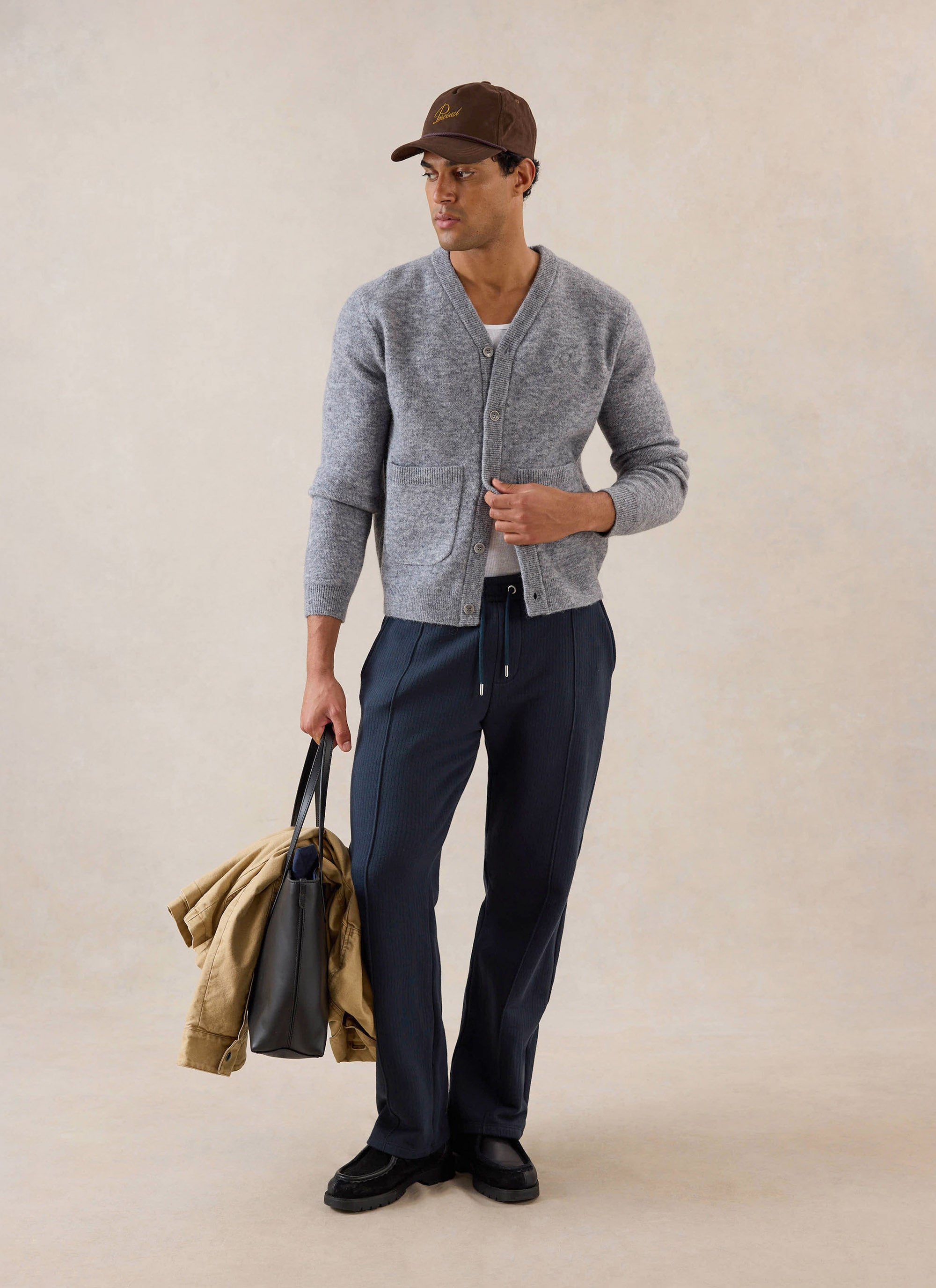 A model wears the grey Percival White Glove Boxy Cardigan, styled with dark trousers, a cap and a tote bag.