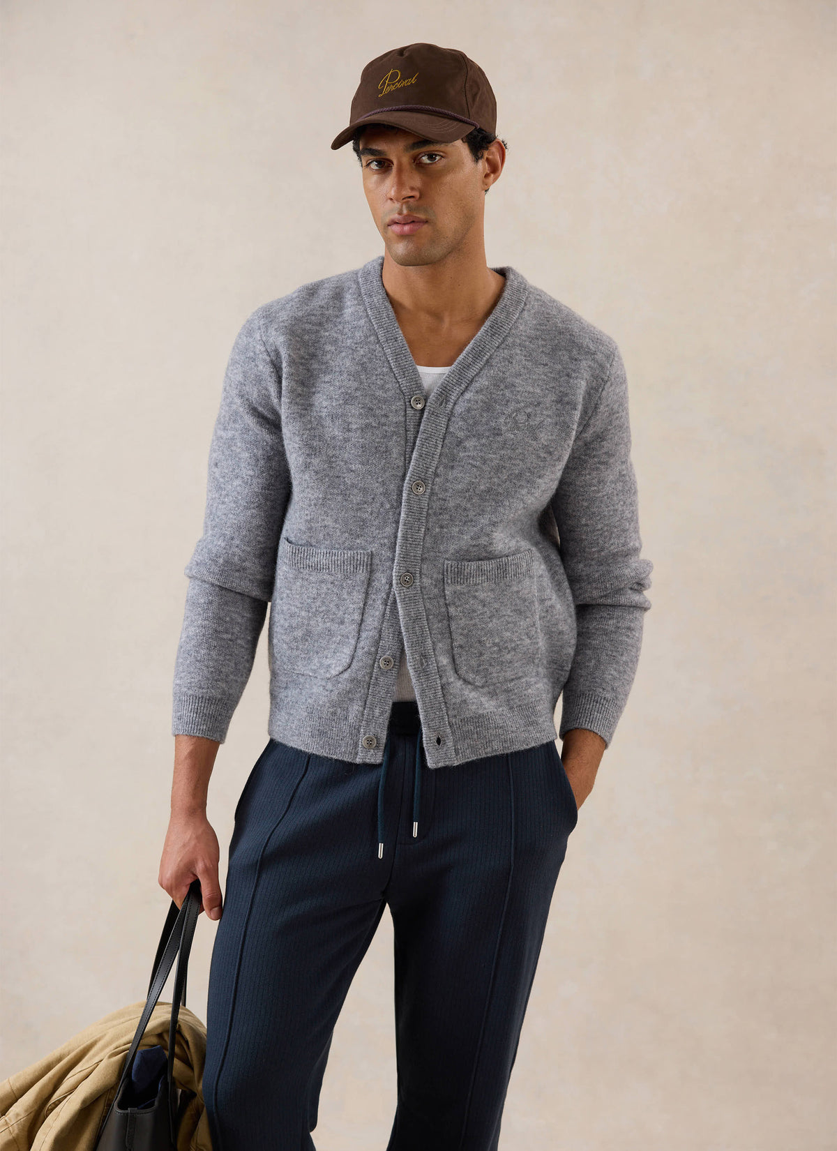 A model wears the grey Percival White Glove Boxy Cardigan, styled with navy trousers, a cap and a bag.
