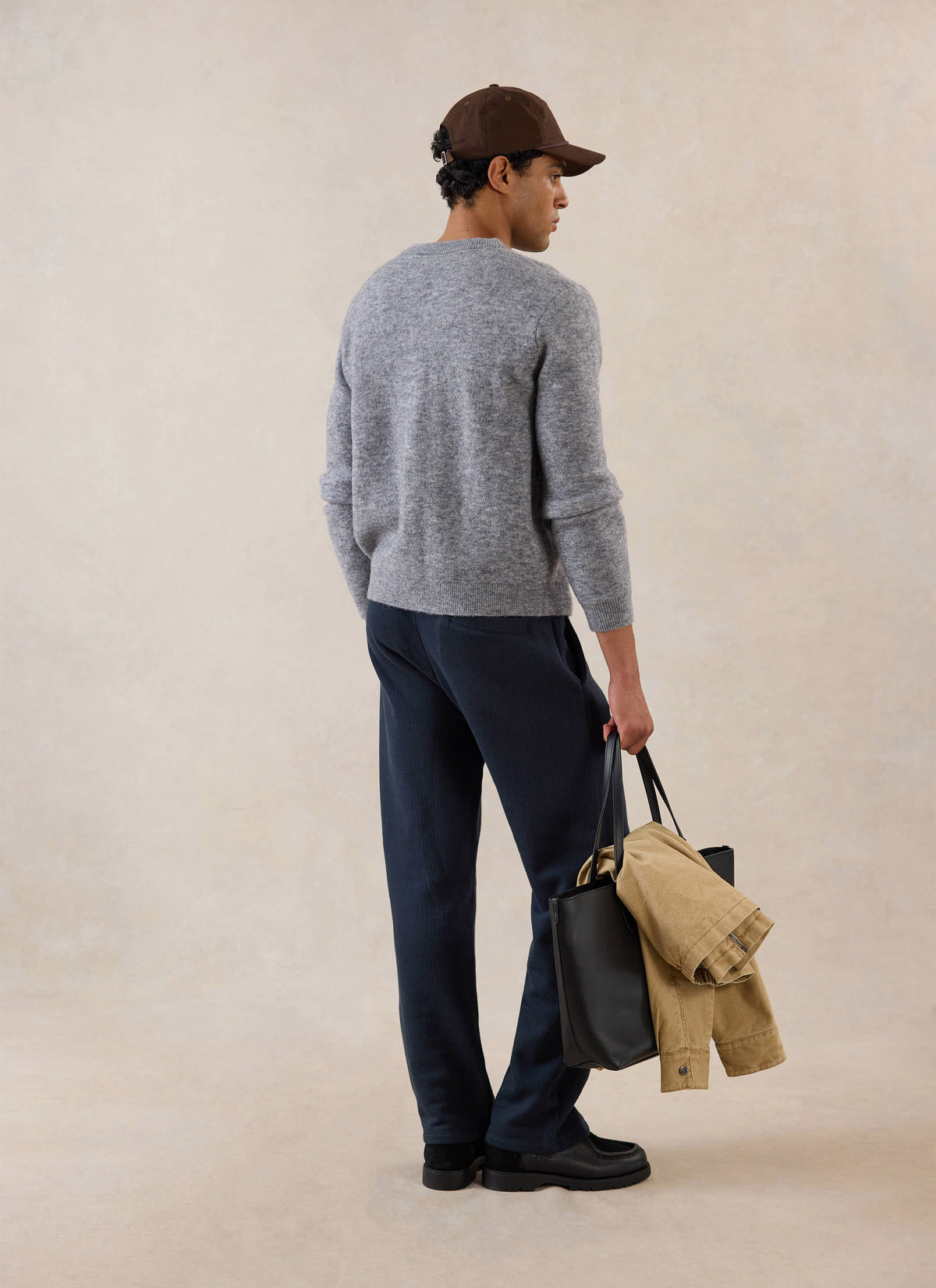 Rear view of a model wearing the grey Percival mohair cardigan, styled with trousers, a cap and a tote bag.