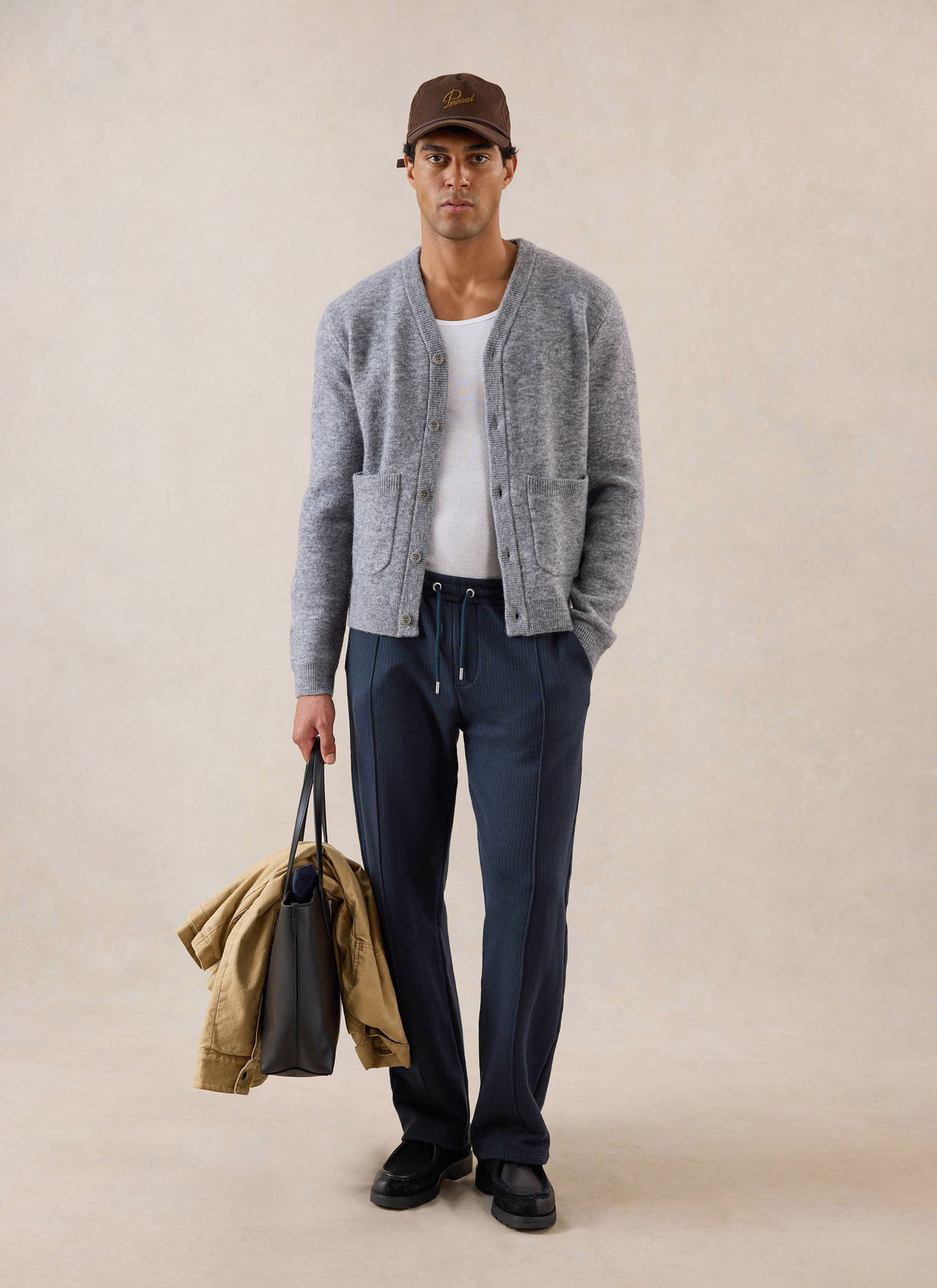 A model wears the grey Percival White Glove Cardigan, styled with blue trousers, a cap, and holding a tote bag in a studio.