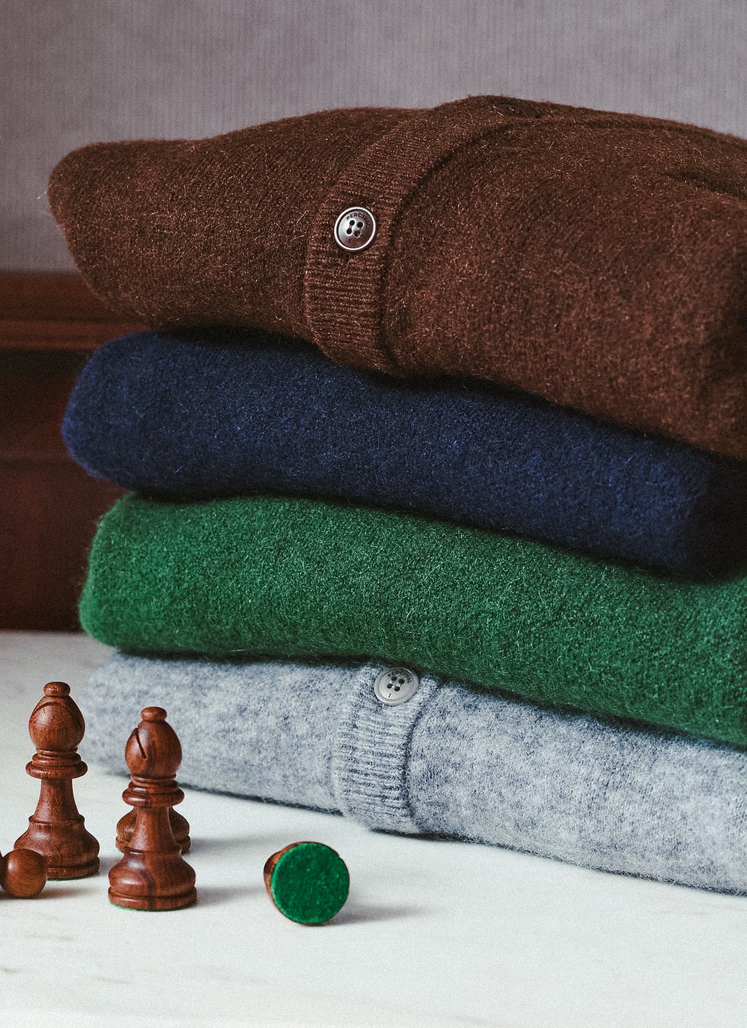 A stack of folded Percival mohair wool cardigans in espresso brown, navy, forest green, and grey, styled with chess pieces.