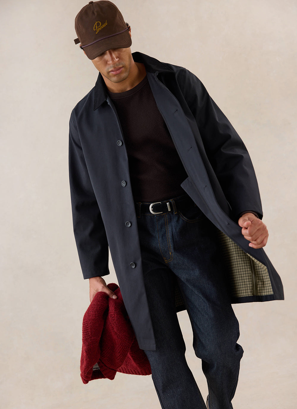 A model wears the black Percival Wilson Long Trench Coat, styled with dark jeans and a cap.
