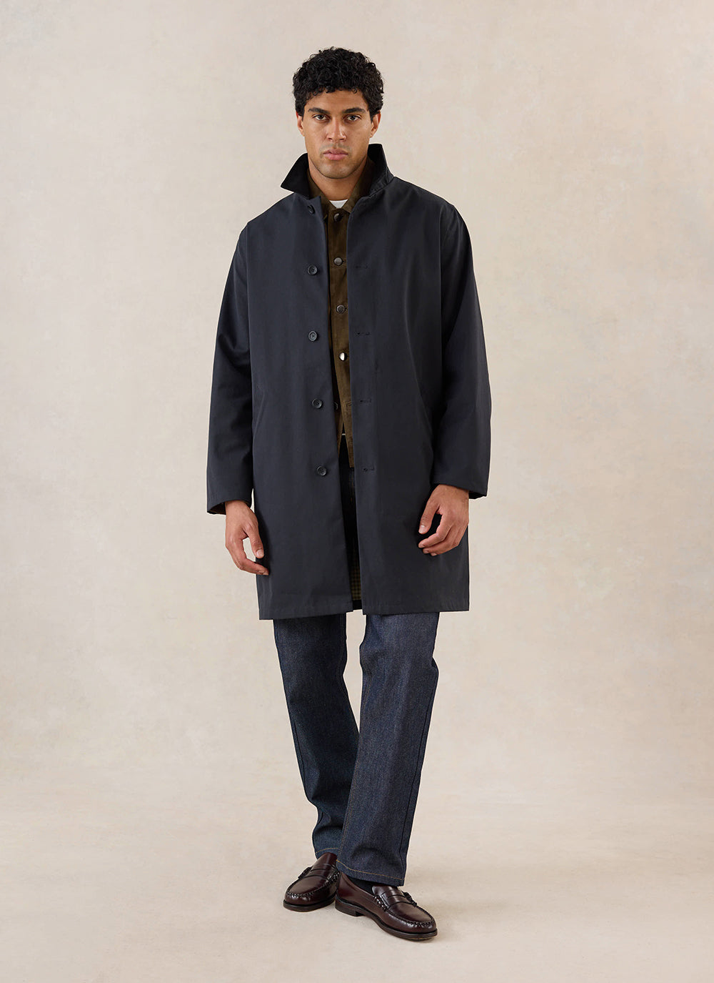 A model wears the black Percival Wilson Long Trench Coat, styled with blue jeans and loafers in a studio shot.