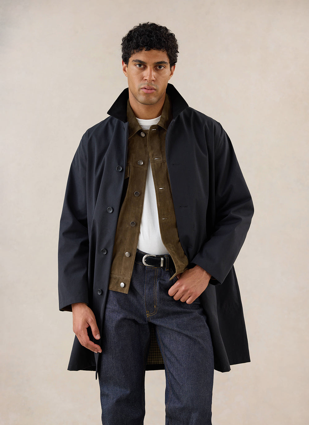 A model wears the black Wilson Long Trench Coat from Percival over an olive overshirt and jeans.