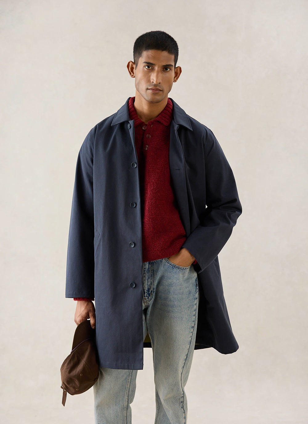 A model wears the midnight Wilson Long Trench Coat from Percival, styled with a red knitted polo and light wash jeans.