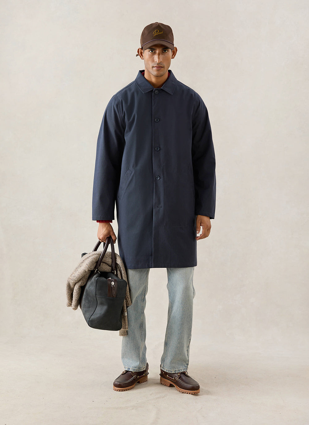 A model wears the midnight blue Percival Wilson Long Trench Coat with a cap, carrying a holdall bag and wearing jeans.