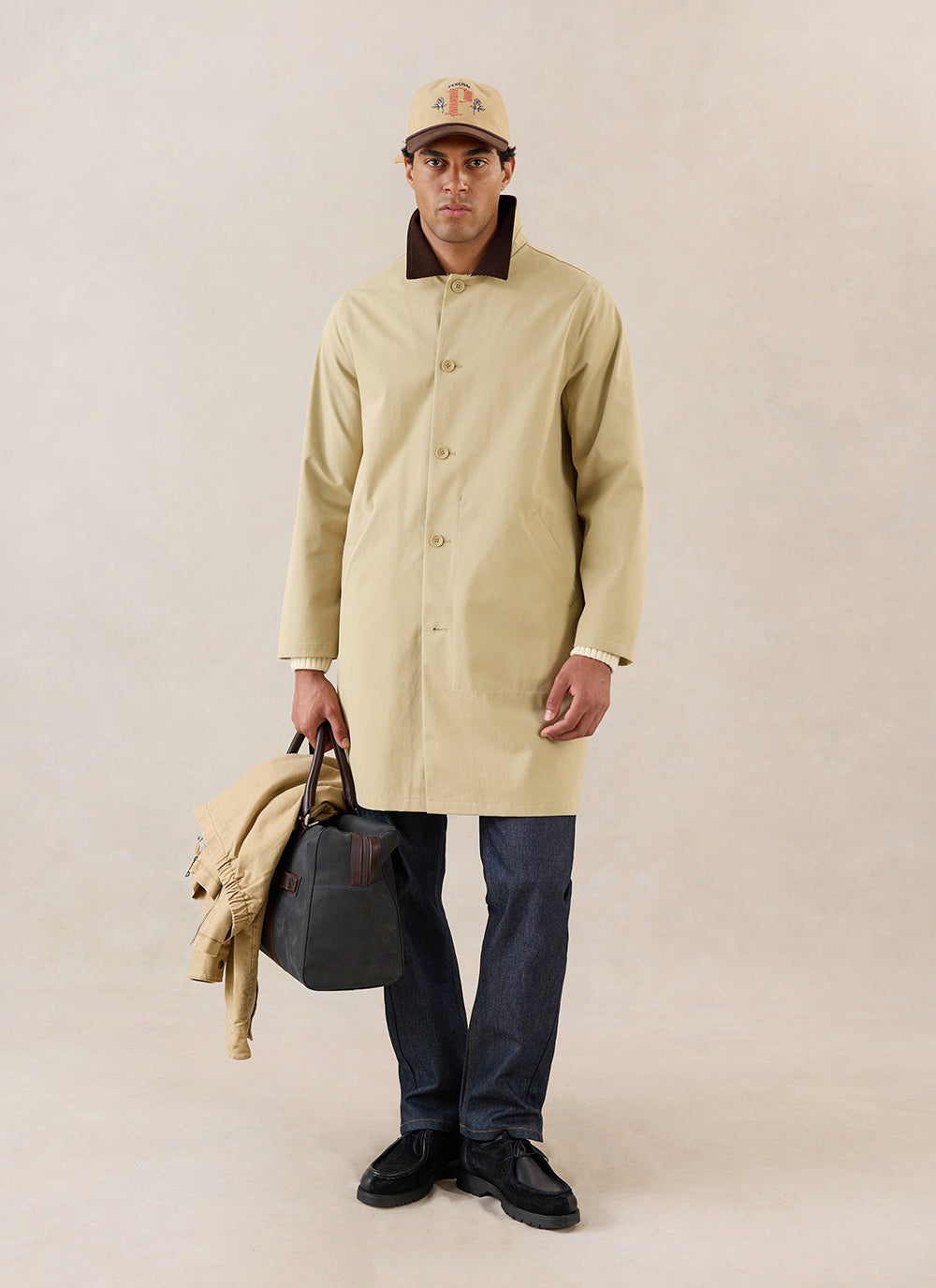 A model wears the tan Percival Wilson Long Trench Coat, styled with a cap, dark trousers, and holding a bag in a studio.