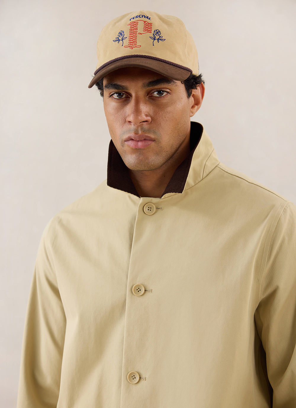 Close-up of a model wearing the tan Percival Wilson Long Trench Coat with a contrasting dark collar, styled with a cap.