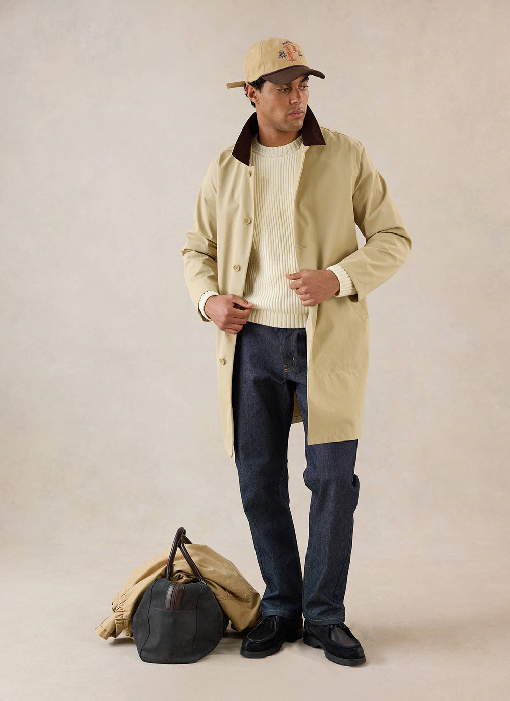 A model wears the tan Percival Wilson Long Trench Coat over a cream knit jumper, dark jeans, cap, and holdall.