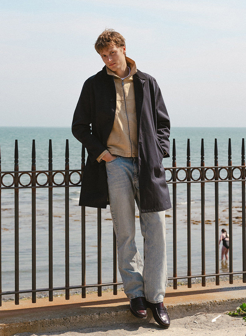 A model wearing a black Percival Wilson trench coat over a beige jacket and jeans outdoors against a coastal backdrop.