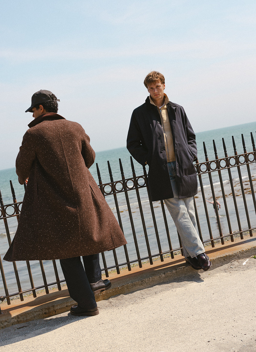 Percival trench coat styled outdoors by the sea, accompanied by a brown fleck coat and jeans.
