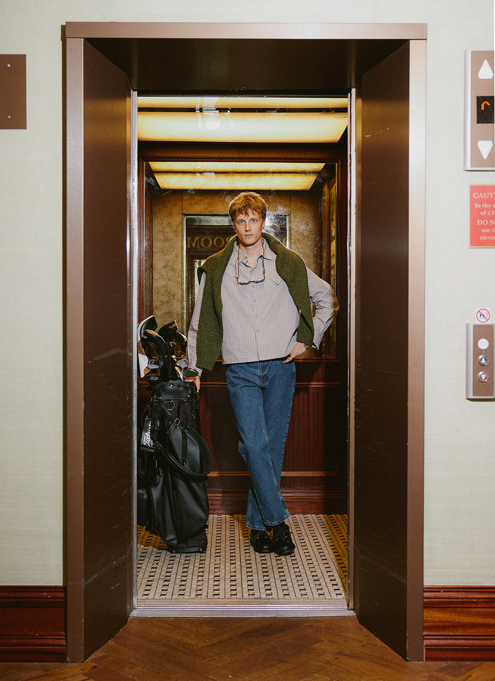 A model in an elevator with a golf bag, wearing the cream Hinsley shirt from Percival layered with a green jumper.