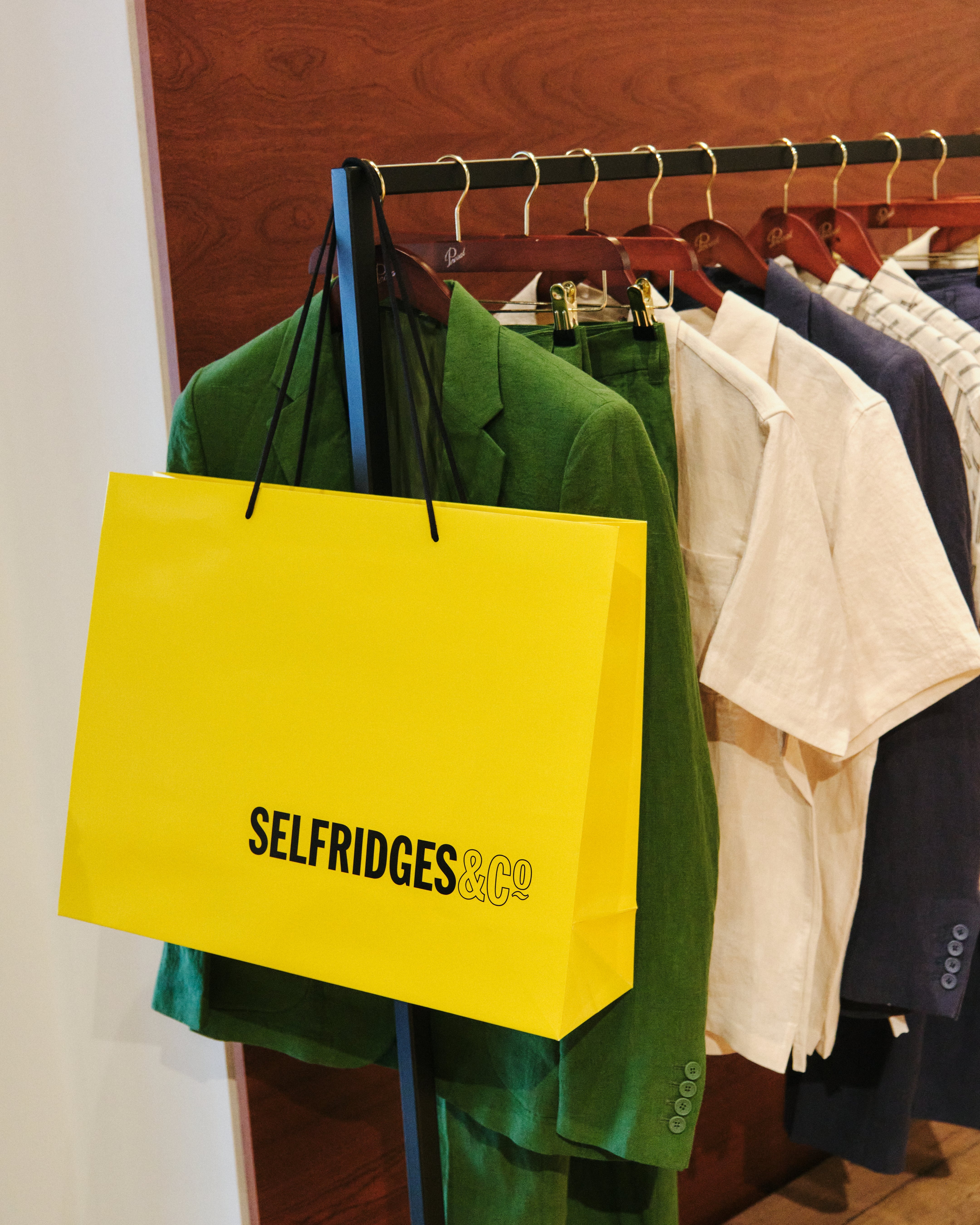 Iconic yellow Selfridges & Co shopping bag placed in front of a rack of clothes at the Percival menswear pop-up store in Birmingham.