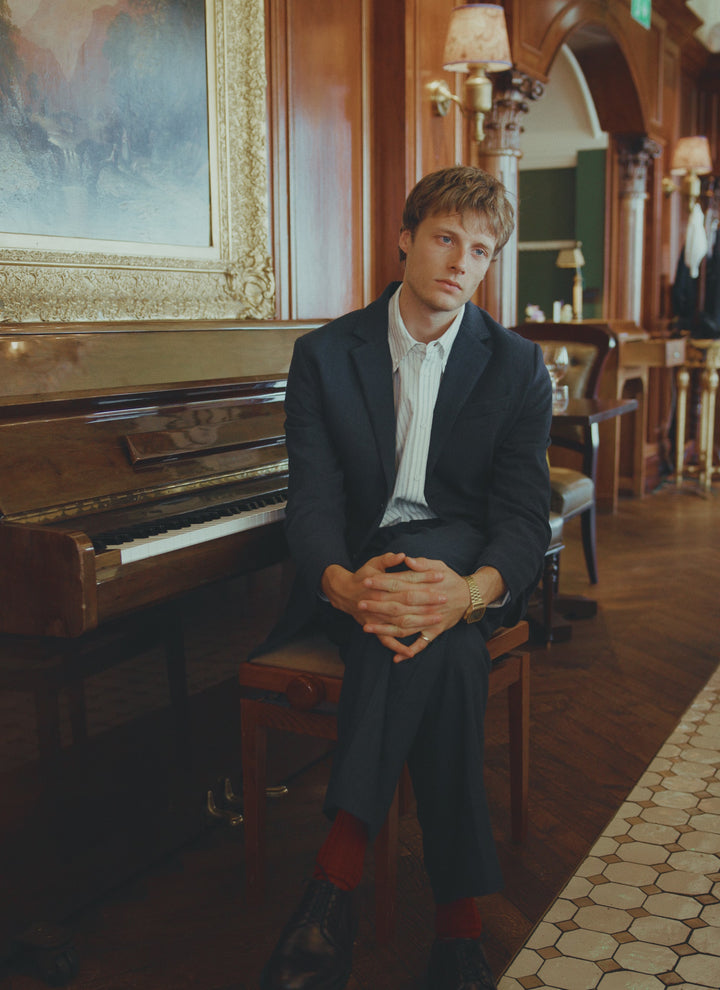A model wears the Percival navy herringbone wool tailored blazer and trousers, sitting at a piano in a wood-panelled room.
