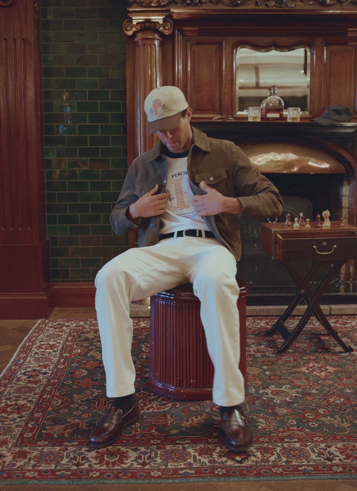 A model wearing the Percival olive suede western jacket seated on a stool, layered over a printed t-shirt and white trousers.