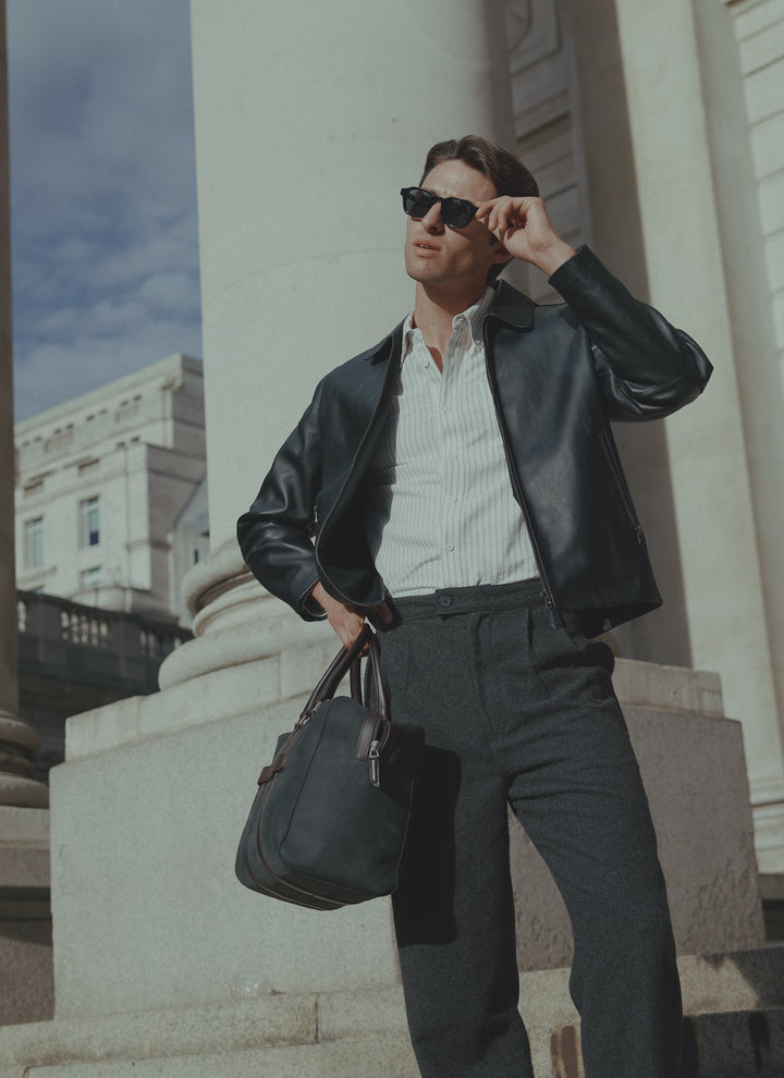 A model wearing the Percival black leather zip jacket, charcoal trousers, and sunglasses outdoors near classic stone pillars.