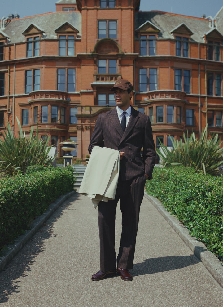 A model wears the Percival Clifton pinstripe pleated trousers in espresso wool, styled with a matching blazer and a cap.