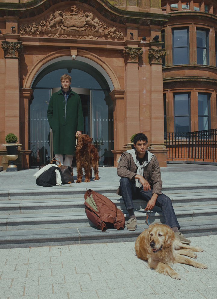 A model wearing the Percival brown herringbone jacket seated on steps with dogs.