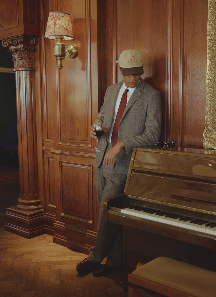 A model wears the grey birdseye wool Percival Blake trousers and matching blazer, styled with a red tie and a cream cap.