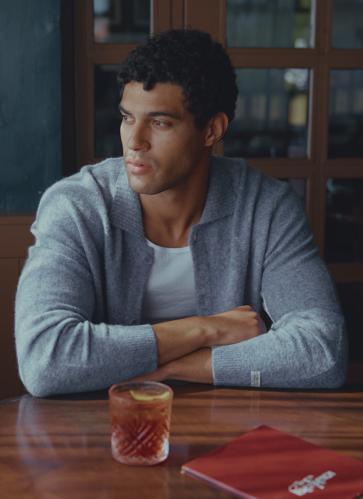 A model wears the heather blue Percival Archive Knit Collared Cardigan while sitting at a table with a drink.