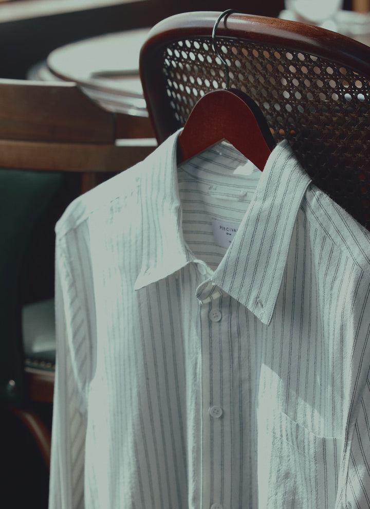 A hand lifts the white Percival Fabian Striped Oxford Shirt hanging on a wooden hanger on a chair.