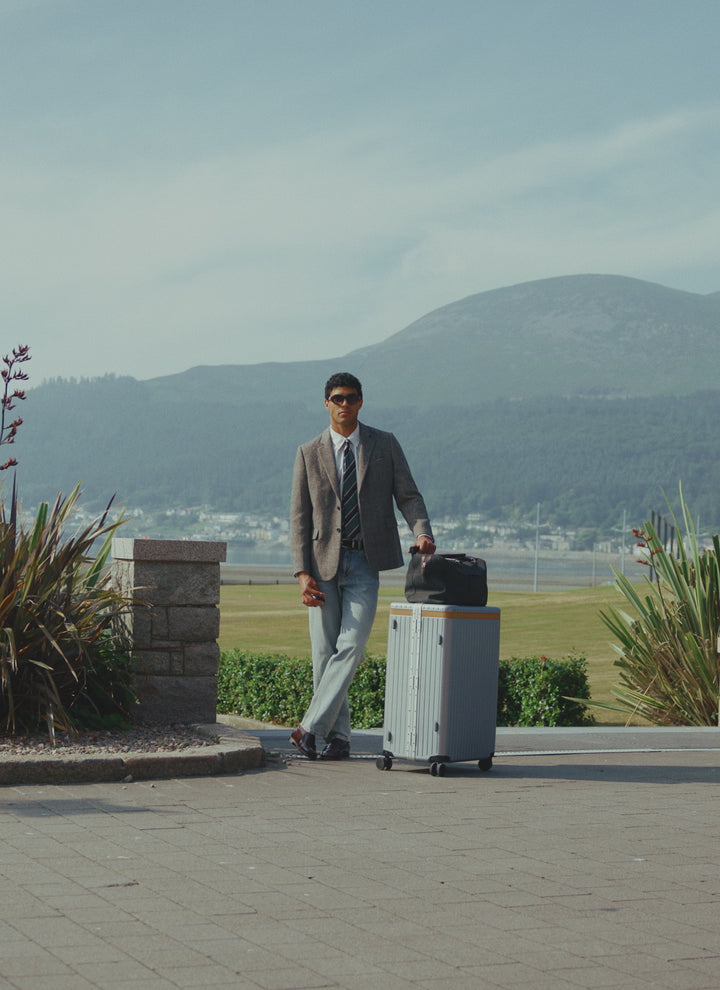 A model wears the Percival grey birdseye wool tailored blazer with jeans and a tie, standing outdoors with a suitcase.