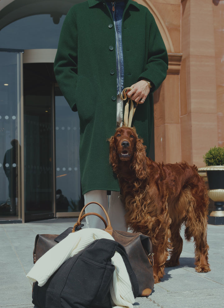 A model wearing the Percival forest green wool overcoat outdoors with a dog, holding a leash next to a leather holdall.