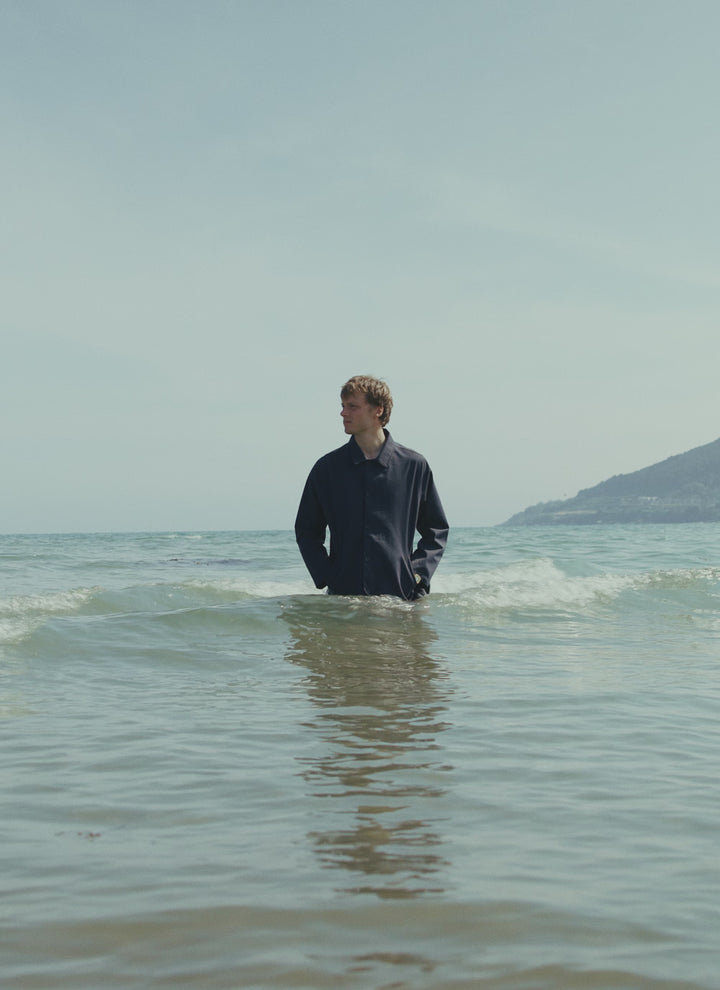 A model standing in the sea wearing the Percival waterproof Sherlock short trench coat in midnight blue.