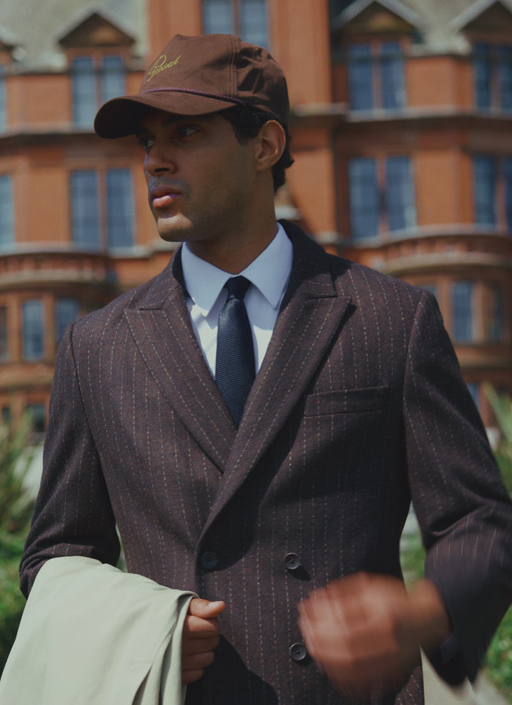 A model wears the Percival espresso pinstripe double-breasted wool blazer, matching cap, and navy grenadine tie outdoors.