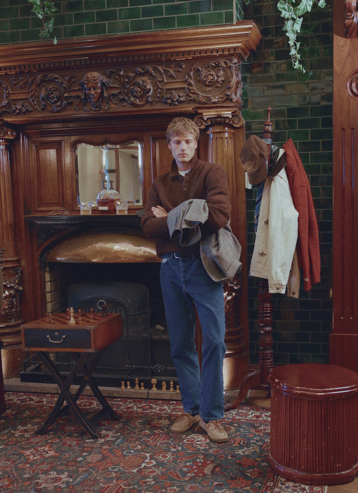 Model wearing the Percival dark brown Shugart knit jumper in alpaca wool blend, styled with jeans and loafers by a fireplace.